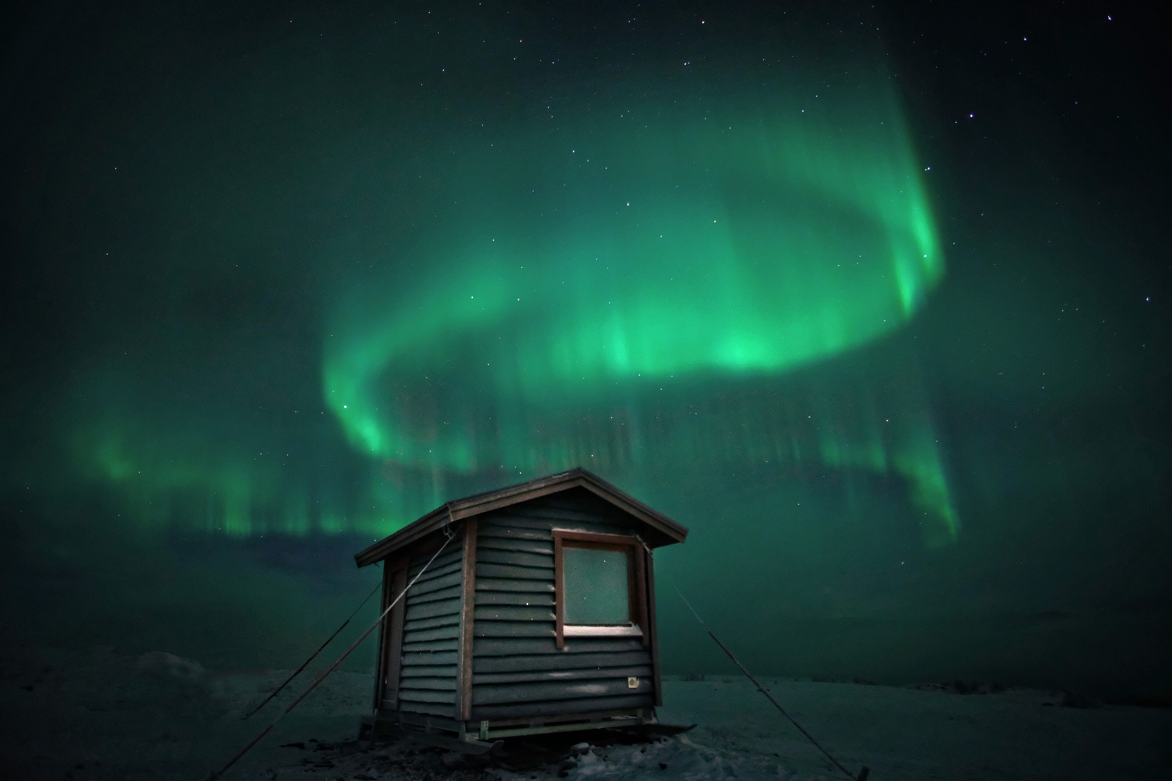 A green aurora arc over a small wooden hut on snow in Swedish Lapland