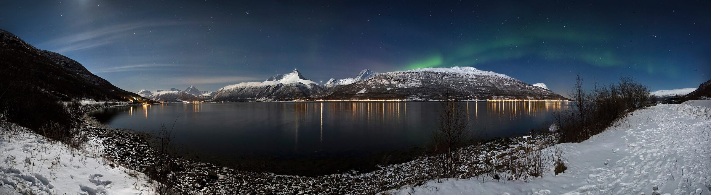 Wide panorama of a fjord and the Lyngen Alps by moonlight with a faint aurora