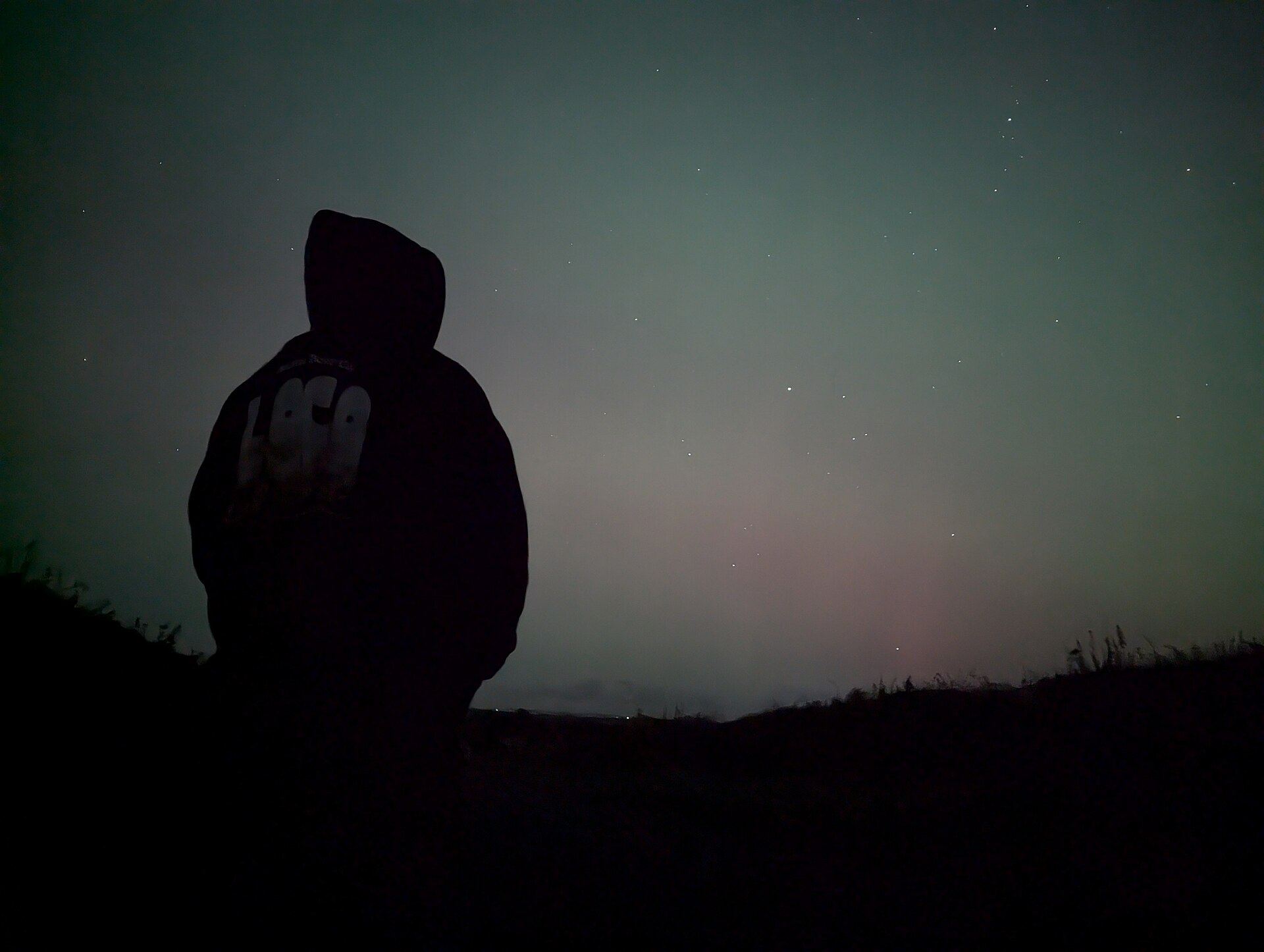 A faint green and pink glow in the night sky above a dark hillside, with a hooded figure in silhouette